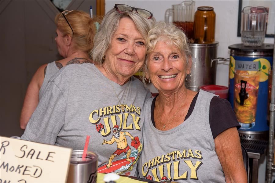 Two women smile together in a brightly lit indoor space. They are wearing matching gray T-shirts.